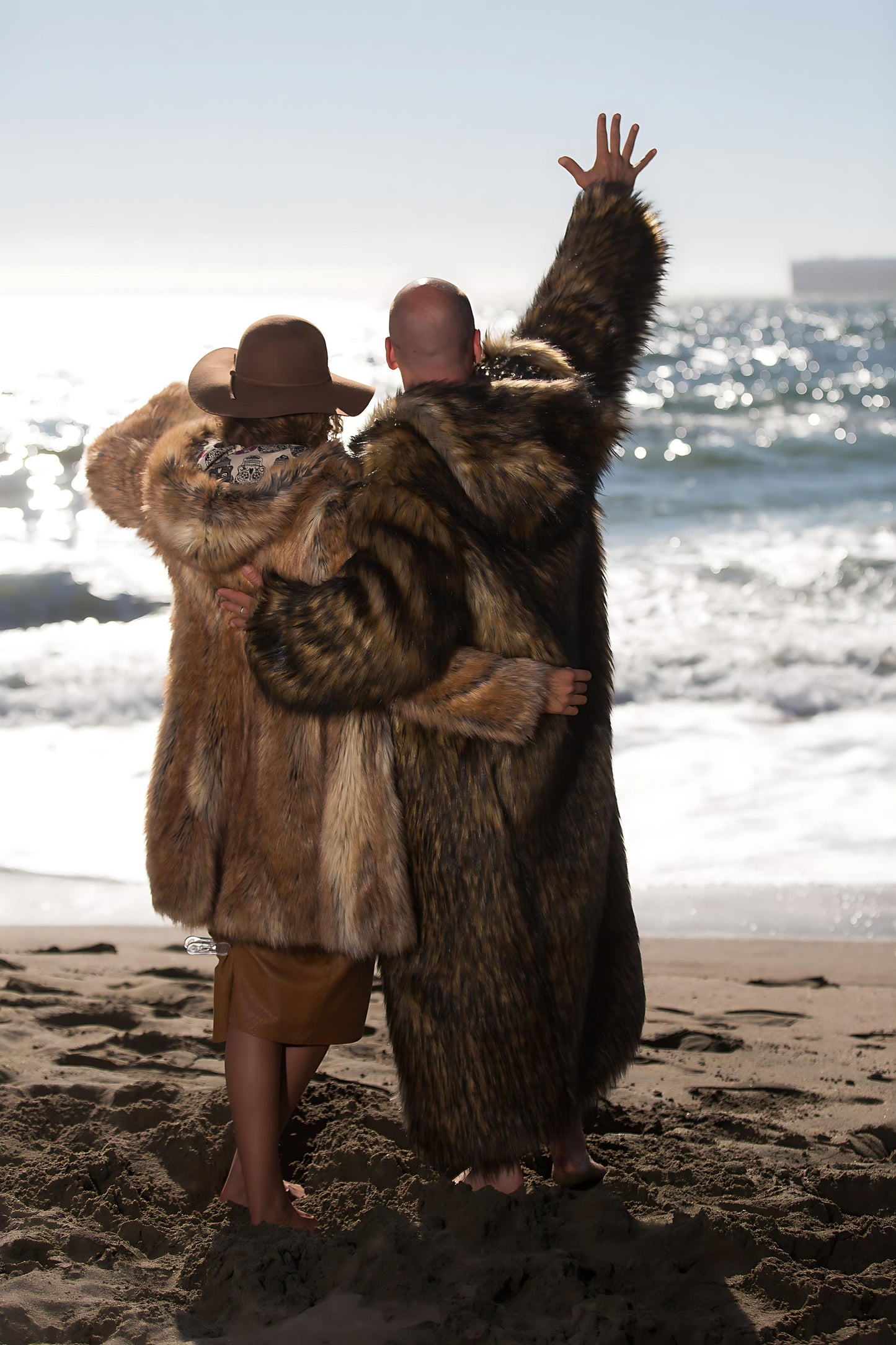 Two kindred spirits in majestic faux fur coats embrace the wild call of the ocean, their hearts in unison with the rhythmic waves, celebrating the unity of man and nature.