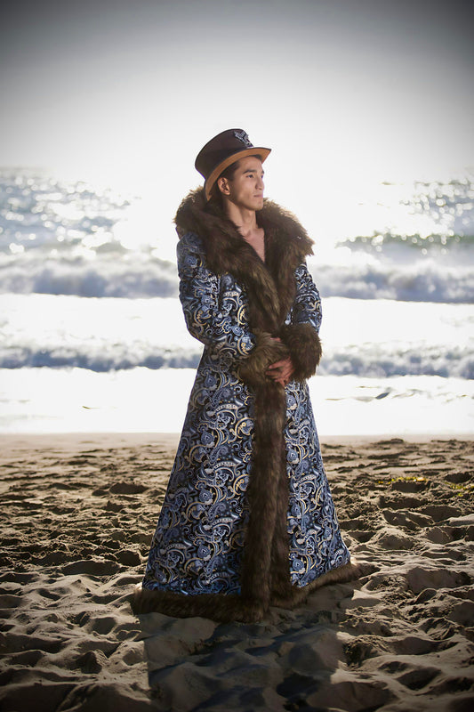 A man stands serenely on the beach at sunset, clad in a Jon Snow BohoCoats faux fur festival coat, its intricate blue and white paisley patterns contrasting with the lush dark fur, embodying a fusion of nature's tranquility and opulent style.